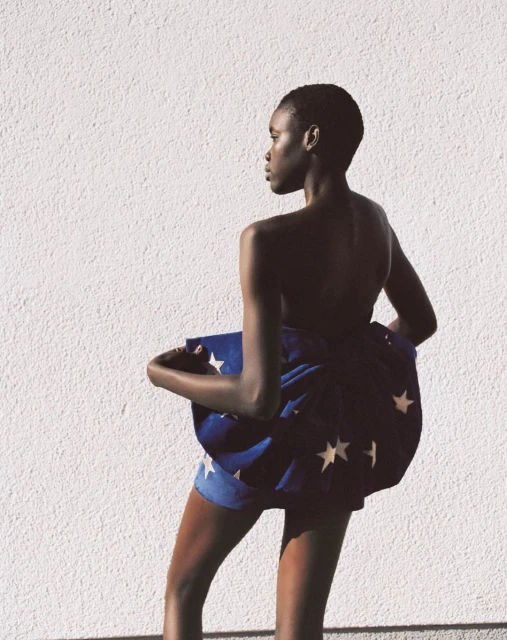 Back view of an African American woman with short hair, draped in blue fabric with white stars against a white wall in natural light.