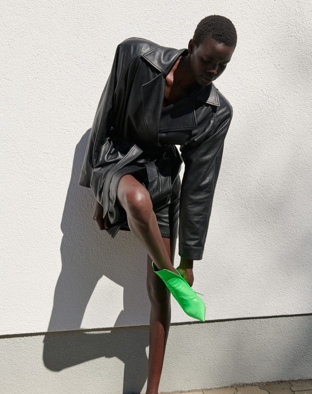 An adult Black woman stands in front of a white wall wearing a black leather coat and putting on a bright green pointed-toe boot.