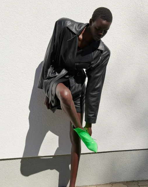 An adult Black woman stands in front of a white wall wearing a black leather coat and putting on a bright green pointed-toe boot.