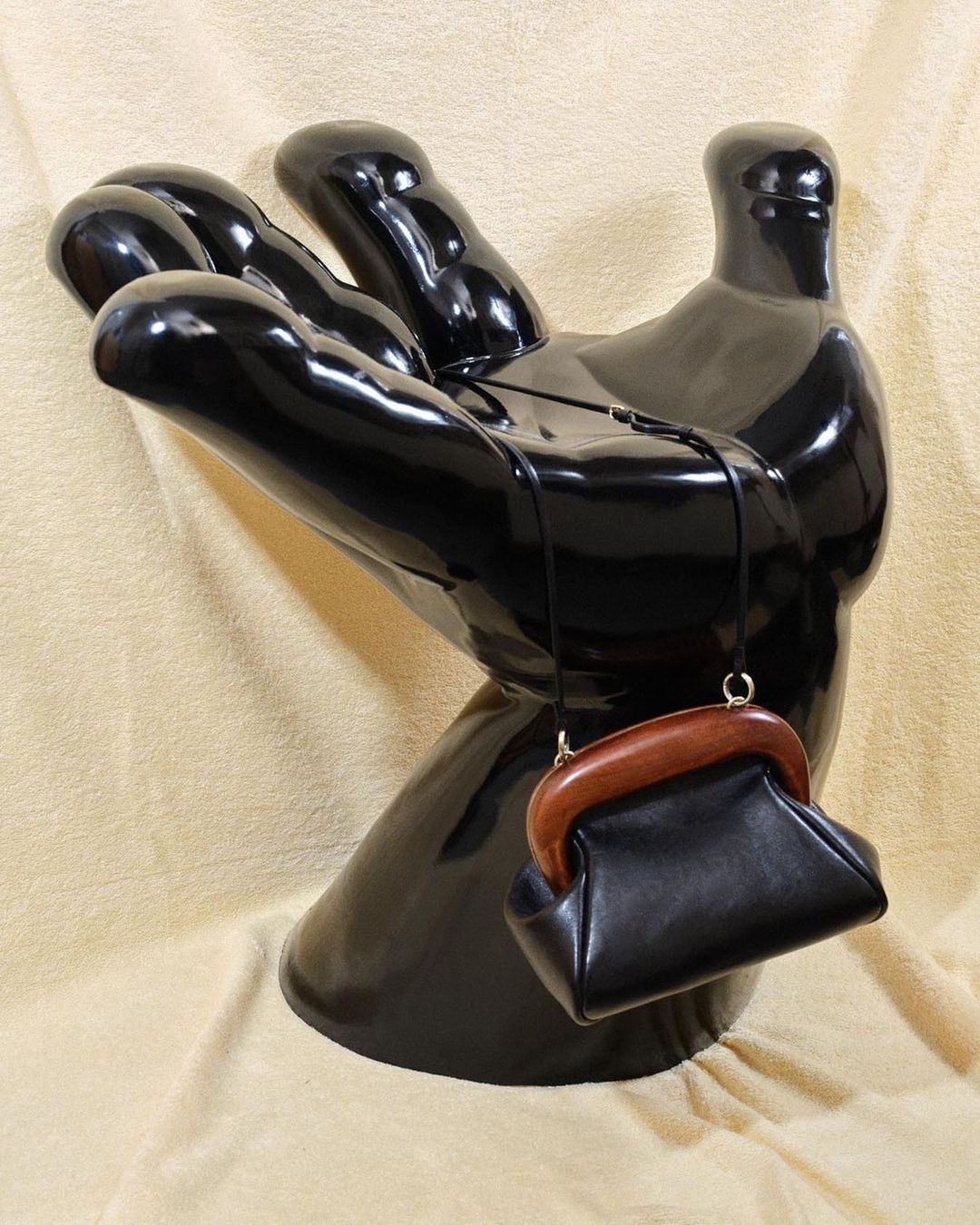 Black purse with wood handle on a shiny black mannequin hand against a beige fabric backdrop. Fashion accessory close-up.