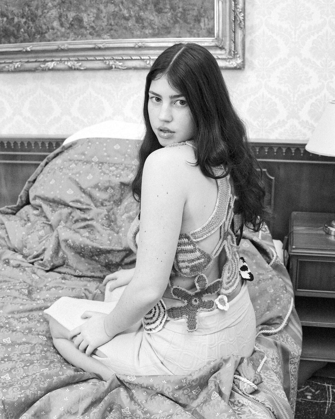 Black and white portrait of a young adult woman in an ornately decorated bedroom, wearing a handmade crocheted top and skirt.