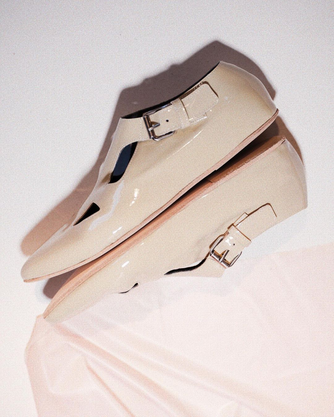 A pair of cream-colored, patent leather Mary Jane shoes with buckle closures, displayed against a neutral backdrop with soft, draped fabric.