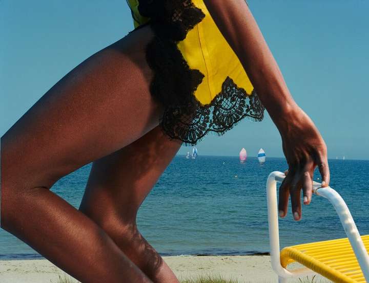 Model poses on a sandy beach near the ocean wearing a yellow and black dress, resting on a chair with white sailboats in the background.