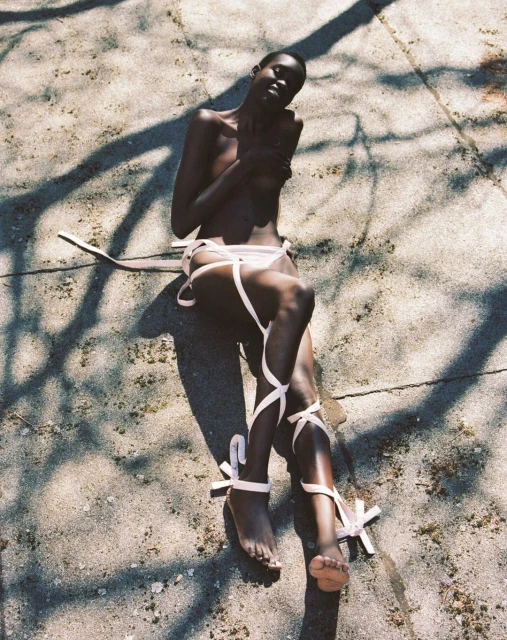Eye-level shot of a nude dark-skinned woman artfully draped in light-pink ribbons, casually reclining on cracked concrete under natural lighting.