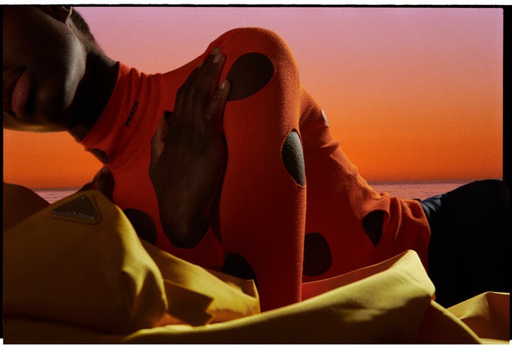 Model in orange patterned top lies on yellow fabric against an orange and purple sunset ocean backdrop, fashion editorial style.