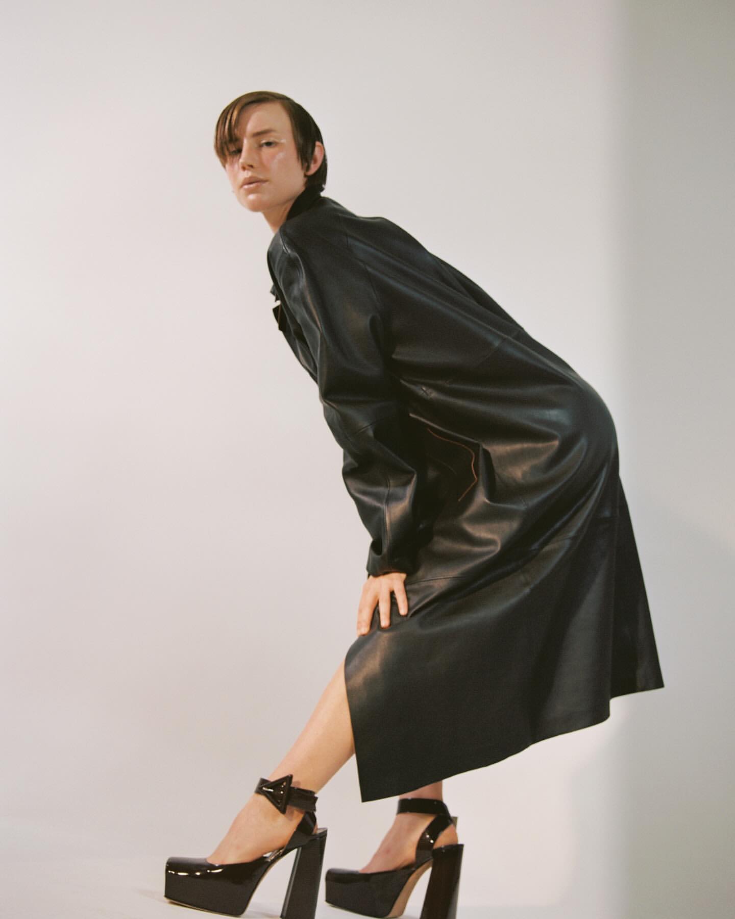 Woman in black patent leather coat and high platform heels posing against a white studio backdrop in a fashion forward style.