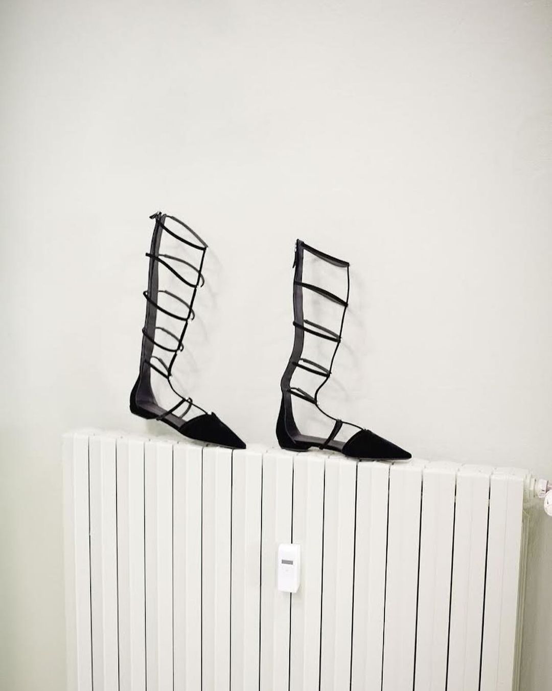 Two black strappy gladiator sandals stand atop a white radiator against a white wall, creating a minimalist fashion footwear product shot.