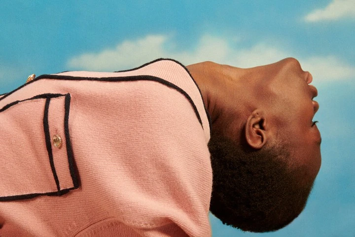 Portrait of a Black adult male with head tilted back wearing a pink knit top with black trim against a blue sky backdrop.