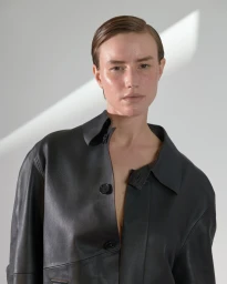 Portrait of a woman with slicked hair wearing a dark leather jacket against a light wall with angled sunlight.