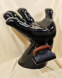 A black leather handbag with a wooden frame rests against a glossy black sculptural hand prop on a textured beige fabric background.