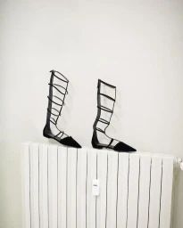 Two black gladiator flats resting on top of a white radiator against a plain wall