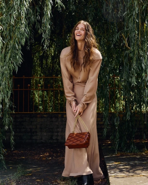 A smiling woman with long brown hair stands outdoors in a flowing tan outfit, holding a brown quilted purse with a gold chain strap.