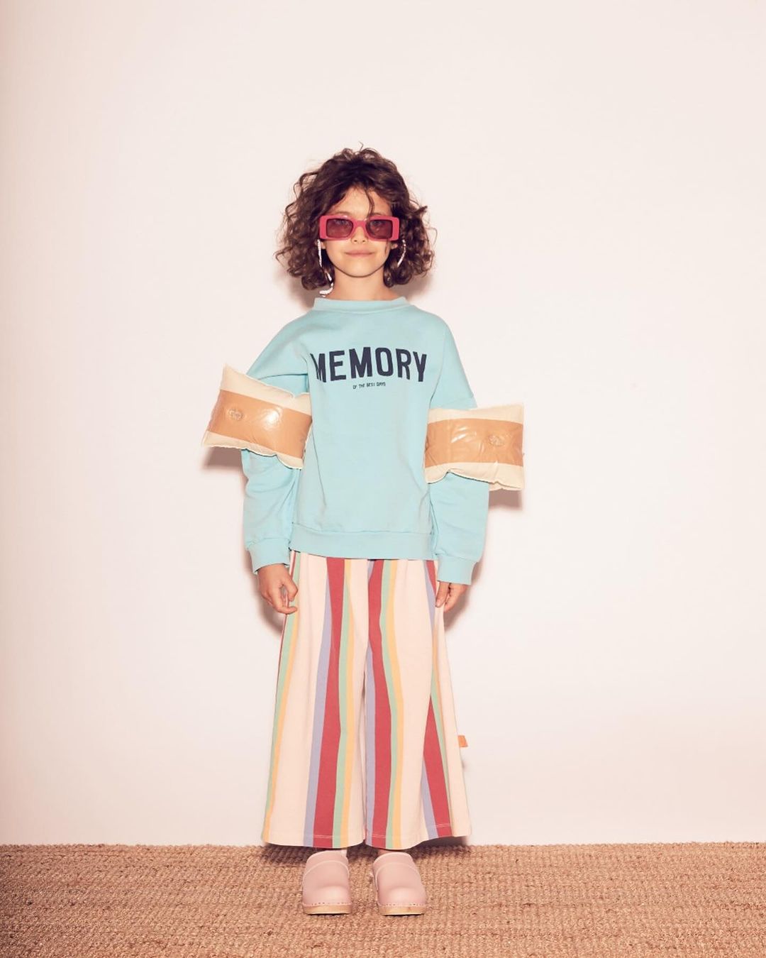 Full shot of child fashion model with curly hair, shades, colorful pants, and aquamarine 'Memory' sweatshirt, posing in studio.