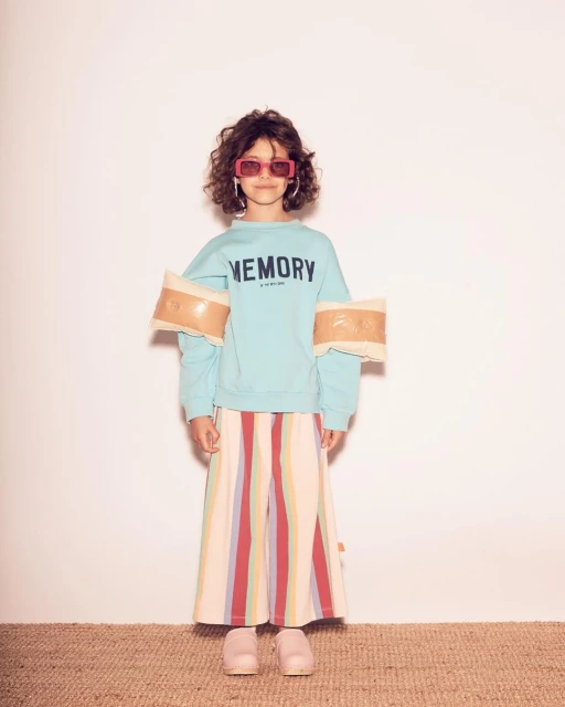 Full shot of child fashion model with curly hair, shades, colorful pants, and aquamarine 'Memory' sweatshirt, posing in studio.