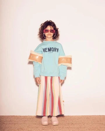 Full shot of child fashion model with curly hair, shades, colorful pants, and aquamarine 'Memory' sweatshirt, posing in studio.