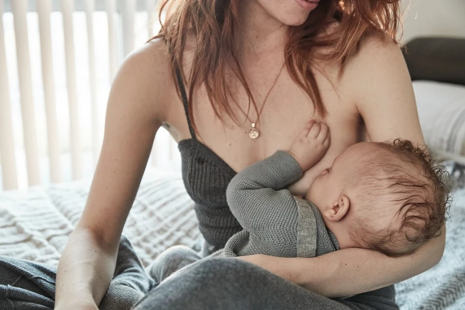 Close-up of a light-skinned woman breastfeeding a baby in an indoor setting with natural light filtering through window blinds.