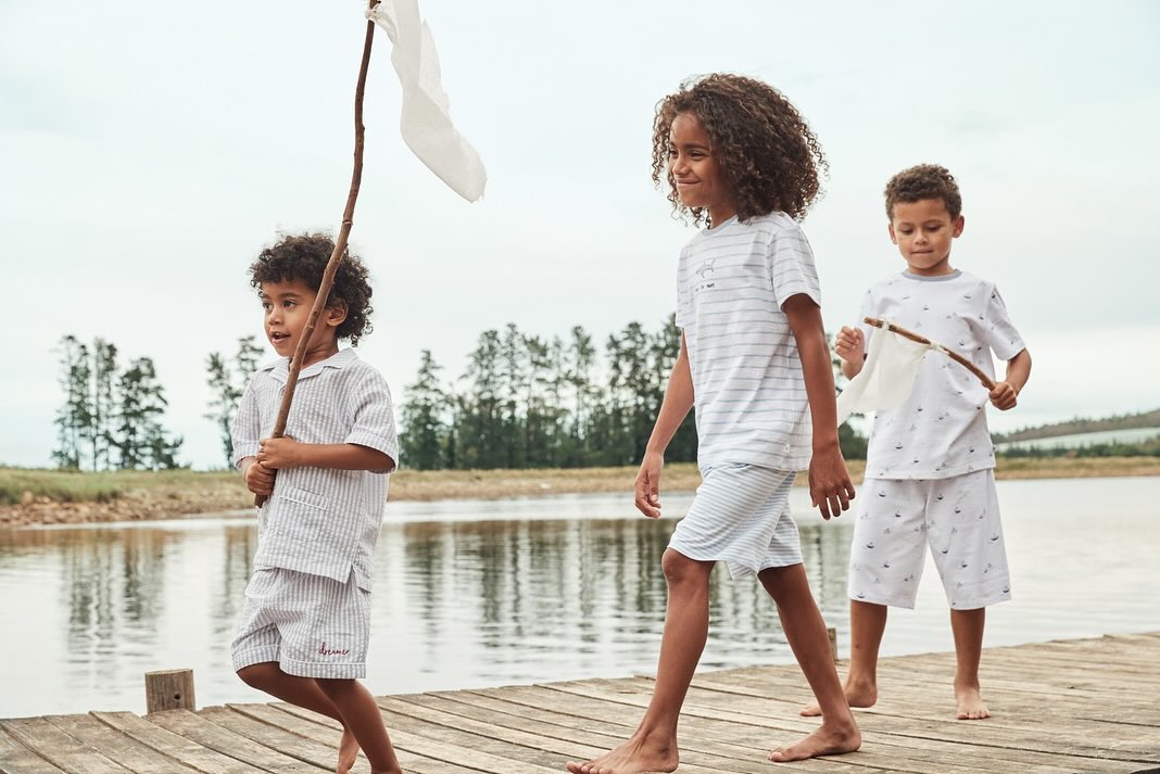 Three children in matching pajamas walk barefoot on a wooden dock by a lake under an overcast sky, each holding a stick.