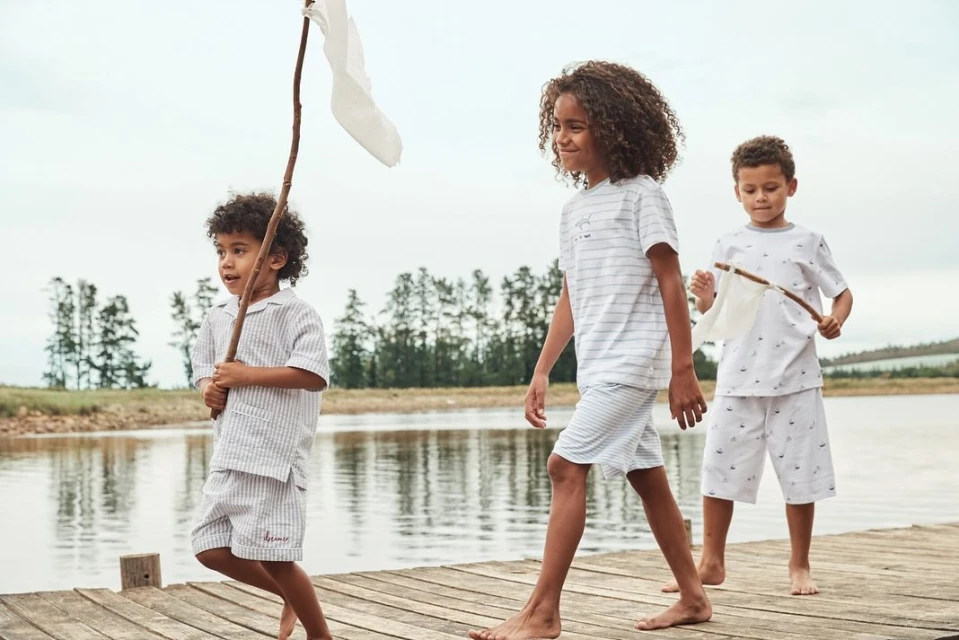 Three children in matching pajamas walk barefoot on a wooden dock by a lake under an overcast sky, each holding a stick.