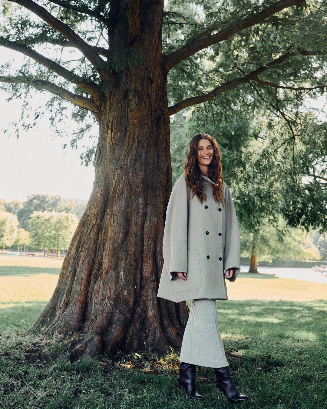 A woman in a coat and ribbed skirt poses next to a large tree in a park setting with a bright sunny sky and grassy field.