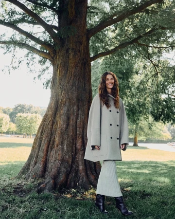 Smiling woman in light grey coat and matching skirt standing by a large tree trunk in a park.