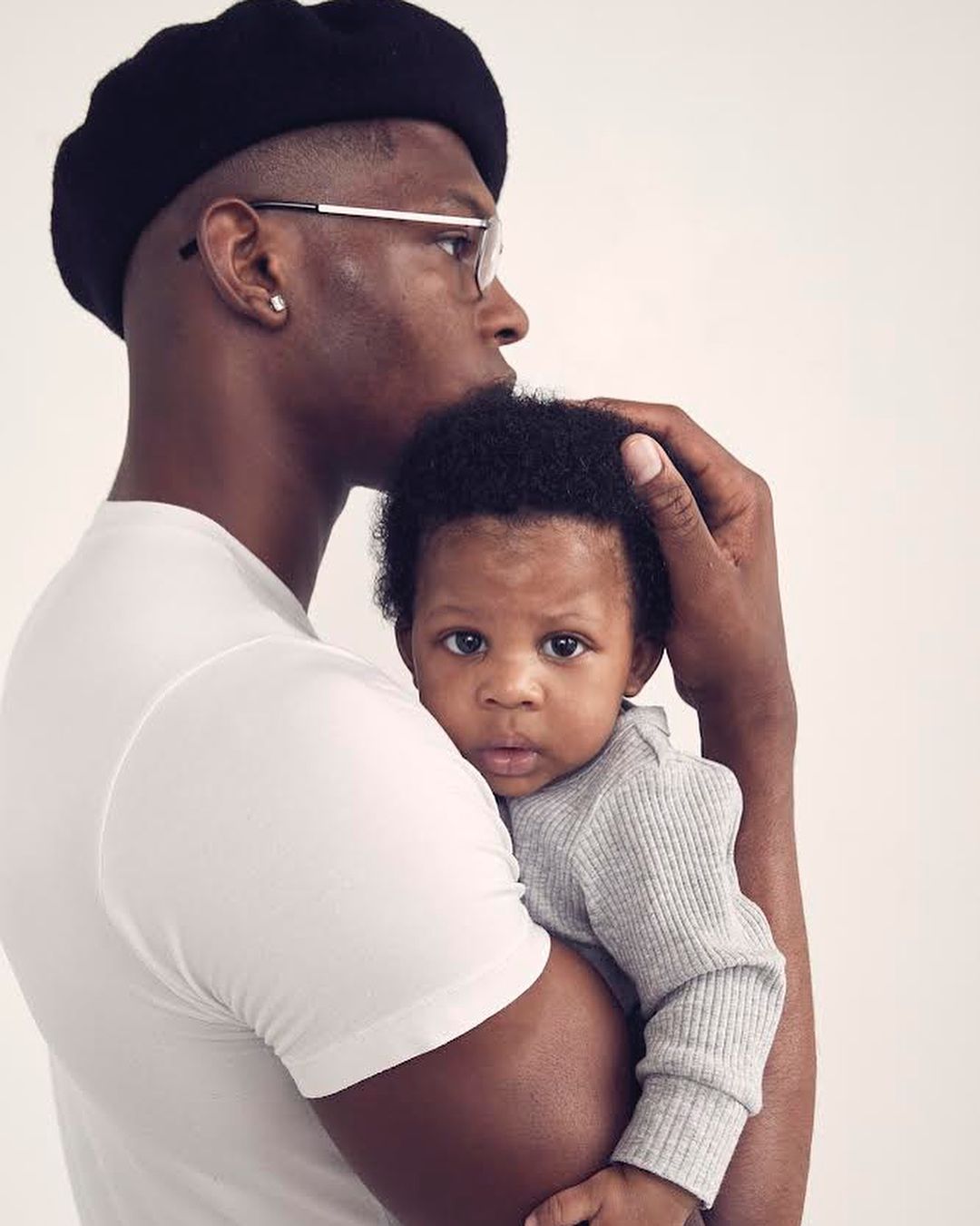 An adult man in a white shirt and black beret holds a baby in a grey top, set against a plain background in a portrait-style shot.