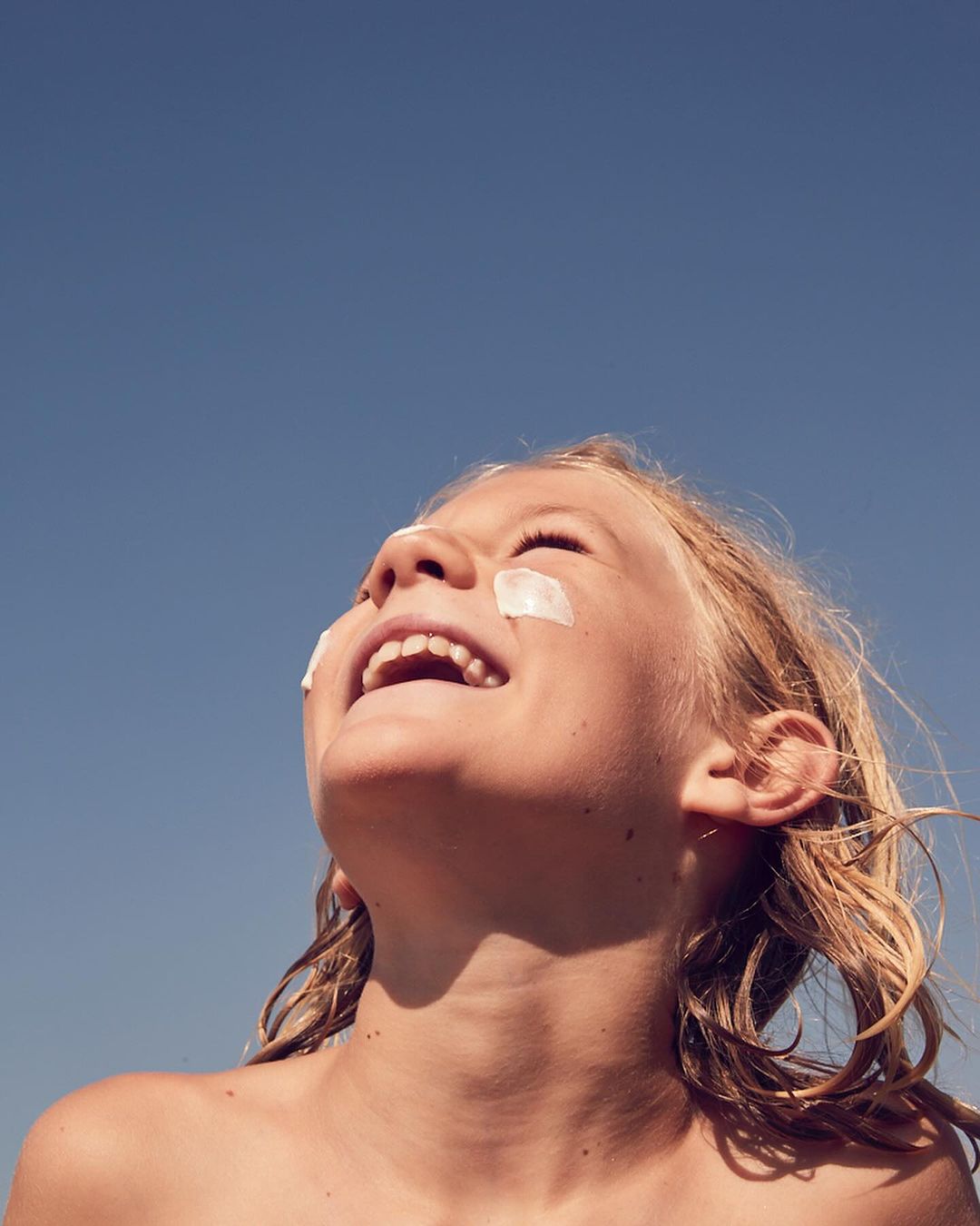 Low-angle shot of a smiling child with white cream dabbed on cheeks against a clear blue sky by Annie Bundfuss
