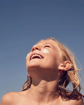 Low-angle shot of a smiling child with white cream dabbed on cheeks against a clear blue sky by Annie Bundfuss