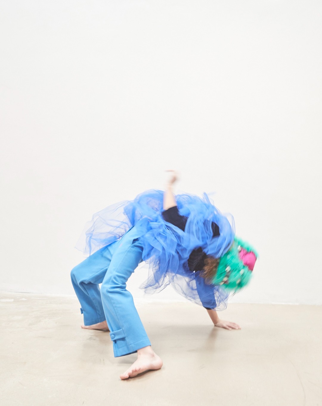Barefoot adult in blue pants and a blue tulle skirt bends over backward, with a green and pink wig, in a studio with a neutral background.