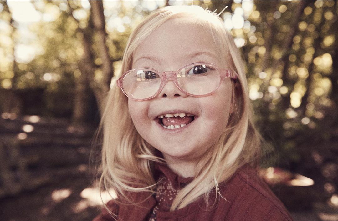 Close-up of a smiling young child with blonde hair and pink glasses in an outdoor setting with blurred trees in the background.