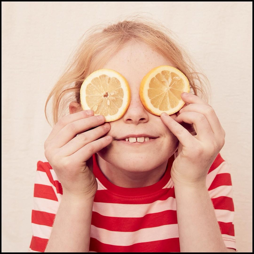 Young girl with blond hair holds sliced citrus fruits over her eyes, striped red and white shirt, and fair skin, smiling indoors.