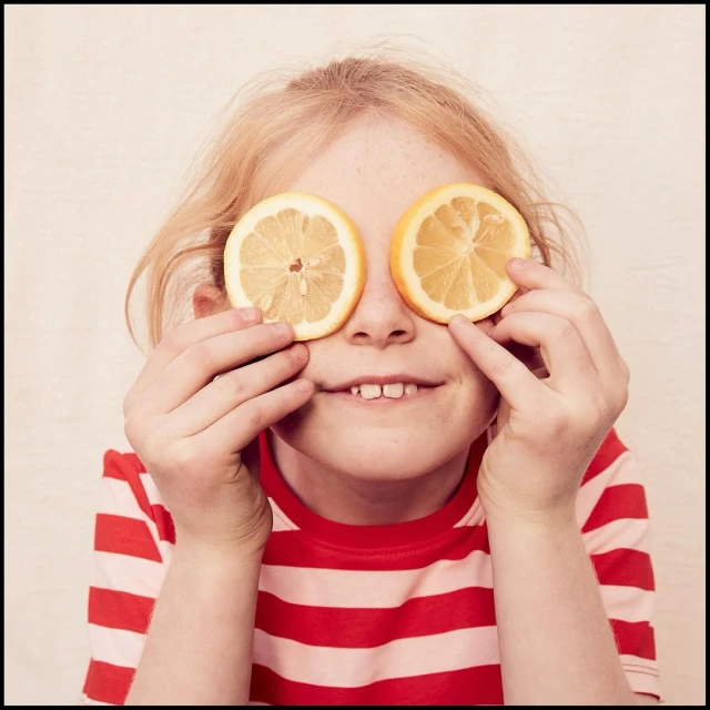 Young girl with blond hair holds sliced citrus fruits over her eyes, striped red and white shirt, and fair skin, smiling indoors.