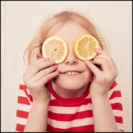 Young girl with blond hair holds sliced citrus fruits over her eyes, striped red and white shirt, and fair skin, smiling indoors.