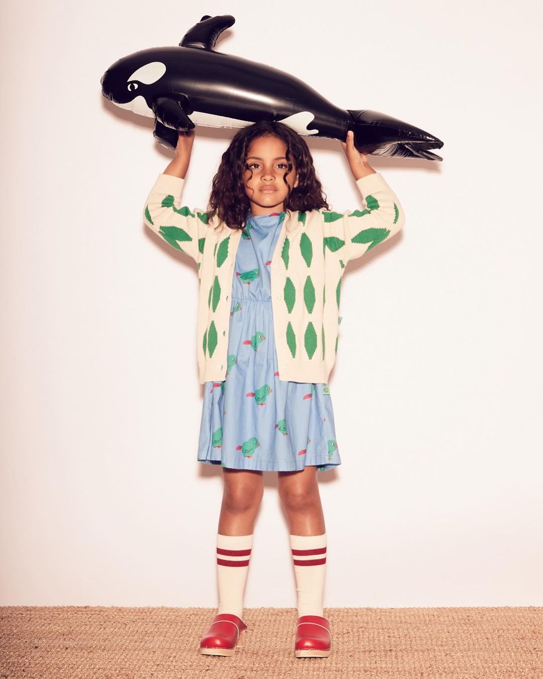 Young girl in patterned dress and cardigan holds inflatable killer whale toy in a studio setting, wearing knee-high socks and red clogs.