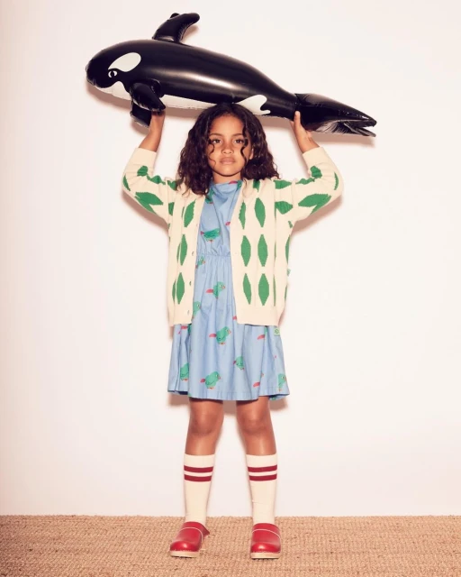 Young girl in patterned dress and cardigan holds inflatable killer whale toy in a studio setting, wearing knee-high socks and red clogs.