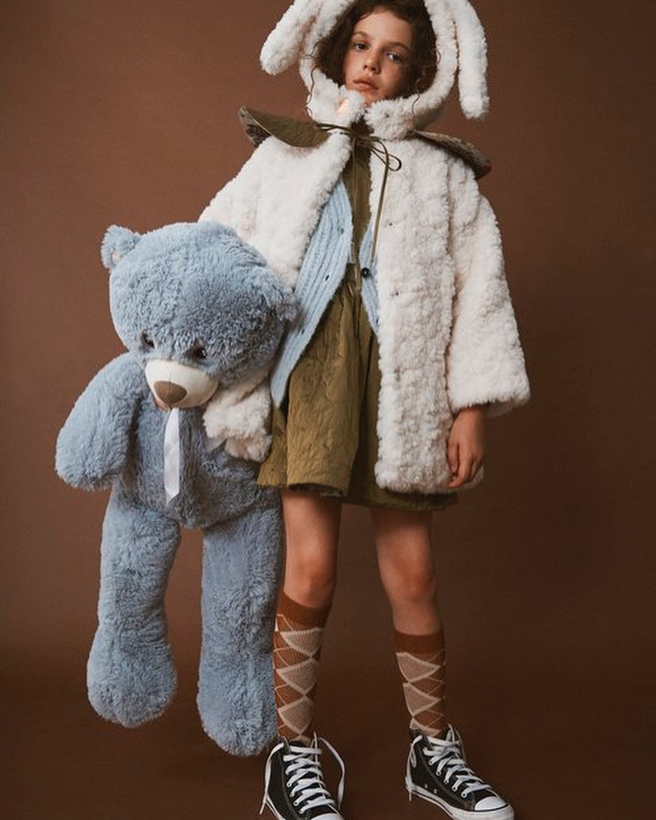 Young child in bunny ears hoody and corduroy clothing holds a blue teddy bear on a brown studio background, children's fashion.
