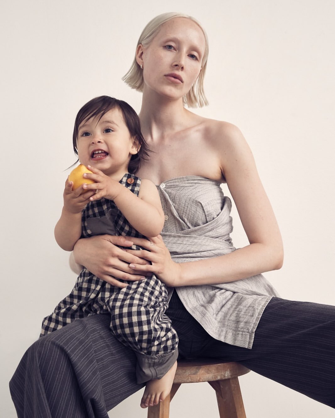 Woman and child sit together in a studio setting. The child is holding an apple. Both are wearing neutral tone clothing.
