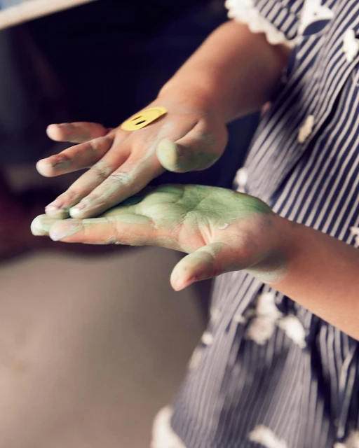 Child's hands covered in green paint, one with a yellow smiley face sticker, and wearing a blue-and-white striped dress, indoors.