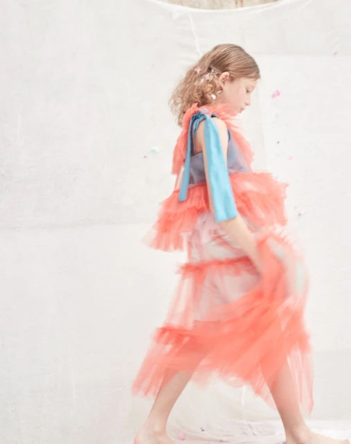 Young girl in a blue and orange tiered tulle dress, walking against a white background in an indoor setting, full body shot.
