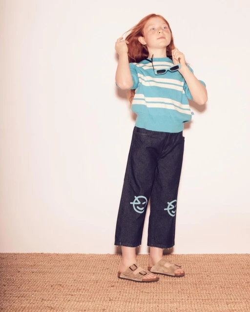 Young girl strikes a pose in a studio, wearing a striped shirt, dark pants, and sandals. She has sunglasses in her hand.