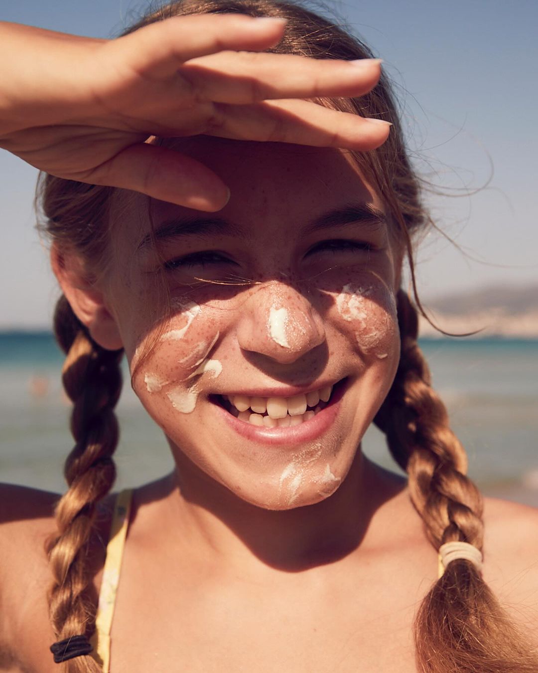 Smiling young girl at the beach shields her eyes with her hand. She has sunscreen on her face and pigtails.