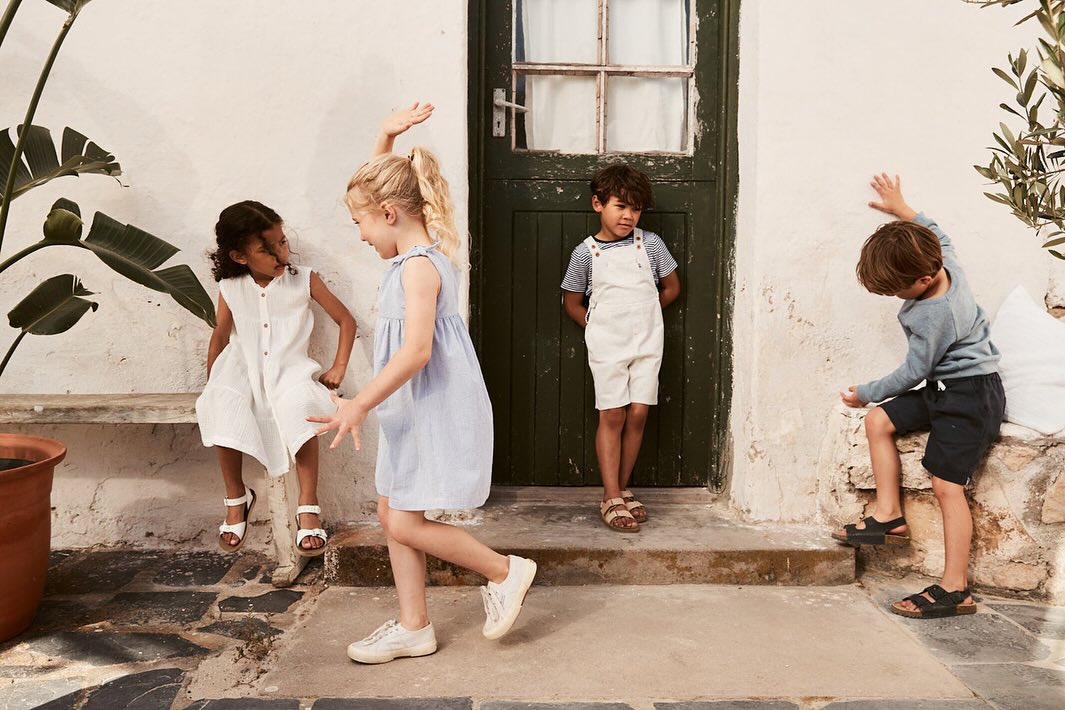 Four kids in casual clothing play near a green door and stone bench outside, bathed in soft daylight, creating a relaxed and playful scene.