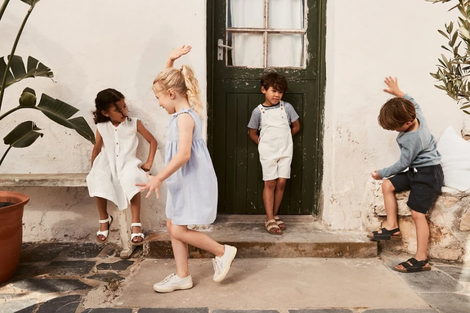 Four kids in casual clothing play near a green door and stone bench outside, bathed in soft daylight, creating a relaxed and playful scene.