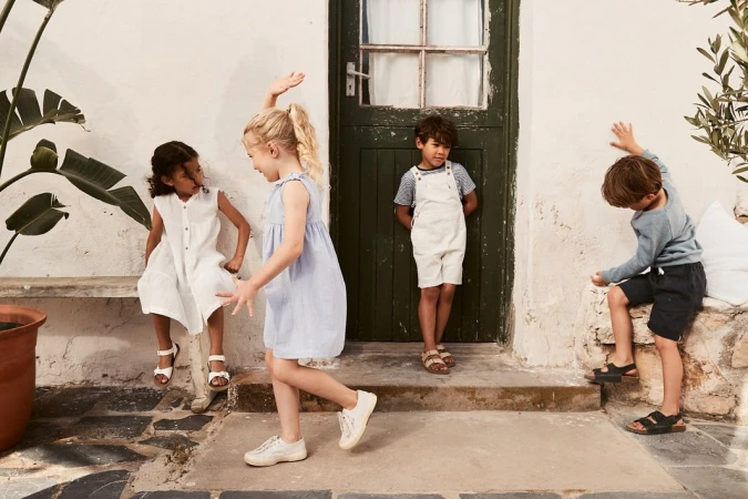 Four kids in casual clothing play near a green door and stone bench outside, bathed in soft daylight, creating a relaxed and playful scene.