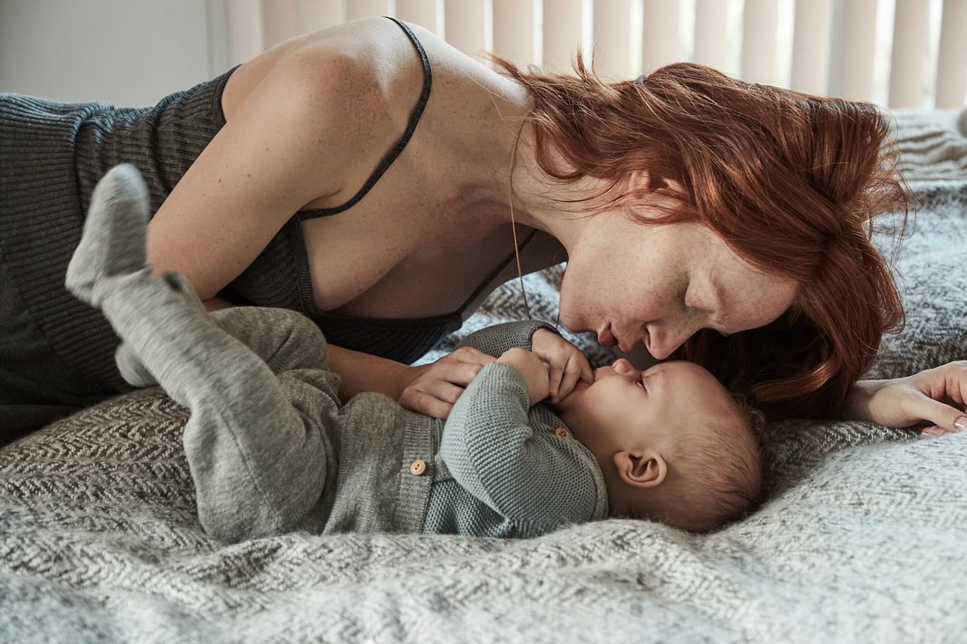 Affectionate indoor portrait of a fair-skinned, red-haired mother lying with her baby dressed in grey on a grey patterned blanket.