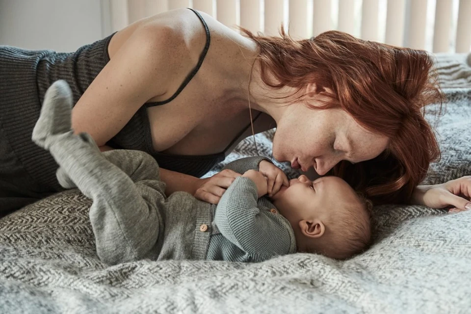 Affectionate indoor portrait of a fair-skinned, red-haired mother lying with her baby dressed in grey on a grey patterned blanket.