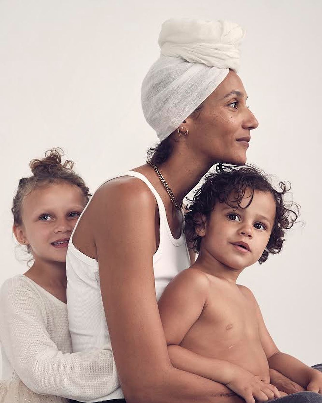 A portrait of a mother with a white headscarf embracing her two children in a studio setting with a plain white background. The image focuses on family.