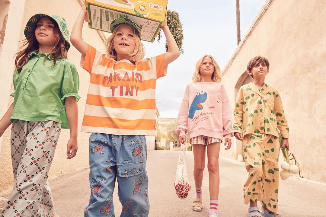 Four kids walk down a sunlit street, wearing colorful clothing and carrying a box and mesh bag, showcasing children's fashion.