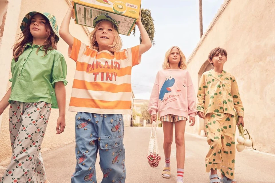 Four kids walk down a sunlit street, wearing colorful clothing and carrying a box and mesh bag, showcasing children's fashion.