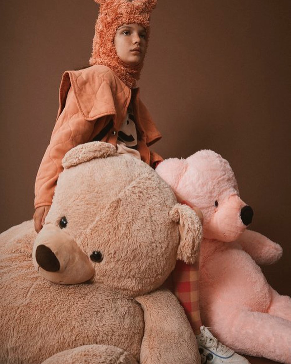 Young person with playful headwear poses with plush teddy bears against a warm brown background in a studio portrait.