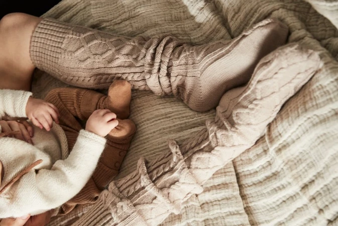 Infant lying on a patterned blanket next to an adult wearing knit socks. Soft, neutral color palette, cozy aesthetic, focus on family and comfort.